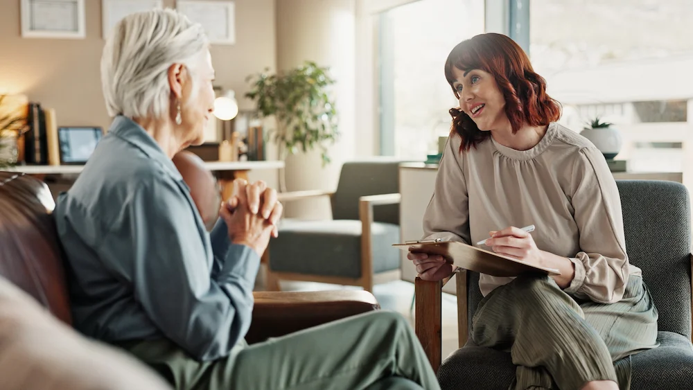 A therapist smiles warmly while holding a clipboard, engaging with an older woman who sits across in a cozy, well-lit office setting, conveying empathy and understanding.