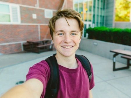 Teenage boy smiling and taking a selfie in a school courtyard. He wears a maroon shirt and backpack. Background shows a brick building and picnic tables.