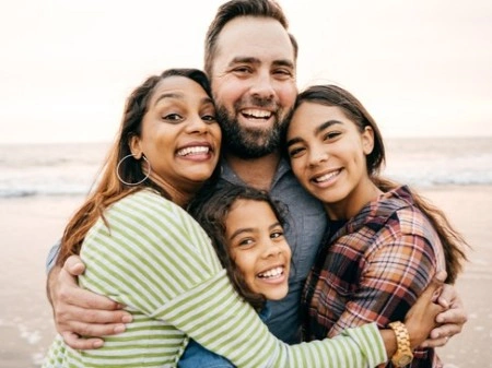 A joyful family of four embraces on a beach at sunset, all smiling broadly. The warm tones and ocean backdrop create a happy, serene atmosphere.