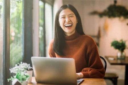 A woman in a rust-colored sweater laughs joyfully while sitting at a wooden table using a laptop. Sunlight streams through a window, creating a warm, cozy atmosphere.