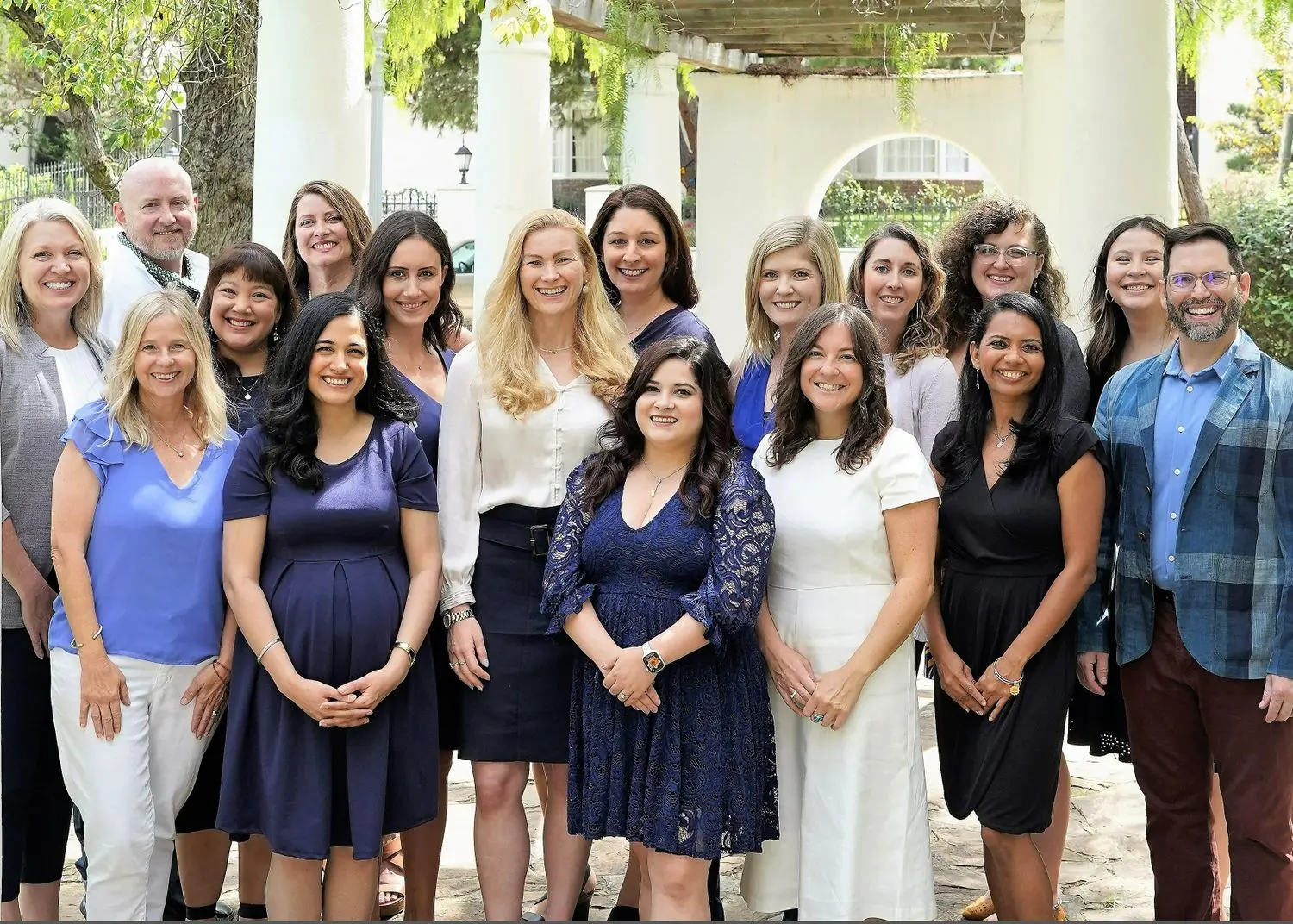 A group of fifteen people, composed of men and women, pose together outdoors. They are smiling, dressed in business casual attire, with greenery and columns in the background, conveying a friendly and professional atmosphere.